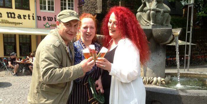 Drei Personen stoßen mit Kölsch am Heinzelmännchenbrunnen in Köln an
